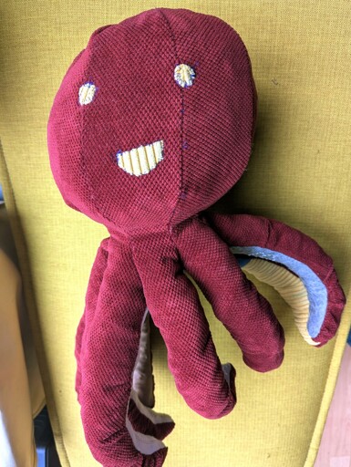 'A high-angle shot of the second octopus plushie resting on a yellow cushion. The red ribbed fabric of its head and tentacles is prominent. It features distinctive yellow stitched facial features: two round eyes and a wide, grinning mouth. One tentacle is curled to the side, revealing a glimpse of the light-colored lining underneath.'