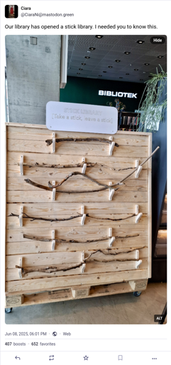 Ciara Nin from mastodon.green has posted "Our library has opened a stick library. I needed you to know this." The alt text says "Dokk1 Aarhus Main Library. inside. Beyond: a white sign on a far wall: Bibliotek. 'Library.' To the right: the edge of a shelf with a plant hanging down. In front of us, about a metre high, a nice wooden display board. It is made of horizontal planks, smooth and light brown, with wooden hooks fashioned into them. Placed on each row of hooks, like valuable swords in a museum, six nice long slim curvy sticks. On top, a sign (in English, even though this is a Danish library): ‘Stick Library. Take a stick. Leave a stick.’ There is no further explanation and nor should we seek one. It is enough that this stick library exists, here, now, before us, for our pleasure, here in this library."