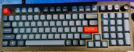 A close-up view of a dark grey Keychron V5 Max mechanical keyboard featuring grey and blue keycaps and a prominent red Esc key and red Enter key. The top right corner includes a programmable rotary encoder knob. The keyboard is backlit. A temporary piece of tape with handwritten text is stuck to the top right keycaps, saying "Print Screen" and "Scroll Lock"