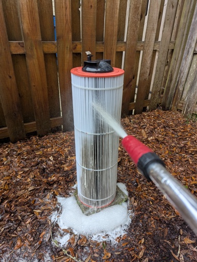 A cylindrical pool filter being blasted with a garden hose jet pressure attachment