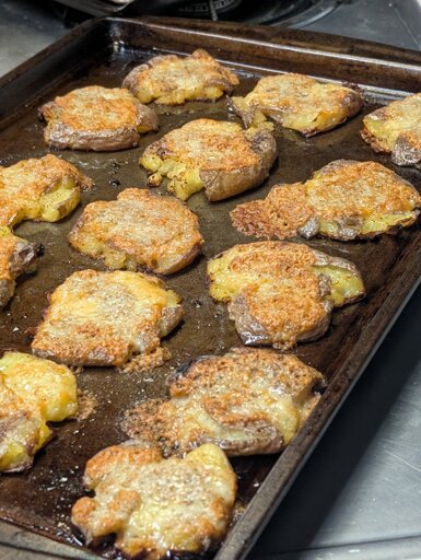 A sheet pan with small flattened potatoes, each covered with a golden lace of melted bubbly cheese.