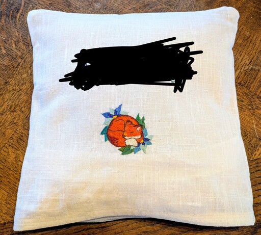 Un coussin blanc avec une broderie d'un renard endormi sur des feuilles. Le nom brodé au dessus est caché.